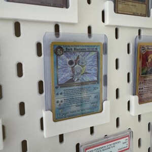 Pu&ograve; includere: Un pannello perforato bianco con diverse carte Pok&eacute;mon esposte in buste di plastica trasparenti. Le carte sono organizzate in file e colonne. Le carte sono in perfette condizioni e presentano una variet&agrave; di diversi personaggi Pok&eacute;mon. Le carte sono etichettate con il nome, il tipo e gli HP del Pok&eacute;mon. Le carte sono anche etichettate con l'anno di rilascio. Le carte sono di diversi colori, tra cui blu, rosso, verde e giallo.