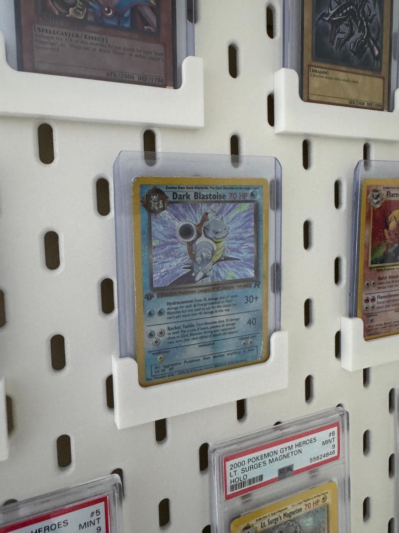 Pu&ograve; includere: Un pannello perforato bianco con diverse carte Pok&eacute;mon esposte in buste di plastica trasparenti. Le carte sono organizzate in file e colonne. Le carte sono in perfette condizioni e presentano una variet&agrave; di diversi personaggi Pok&eacute;mon. Le carte sono etichettate con il nome, il tipo e gli HP del Pok&eacute;mon. Le carte sono anche etichettate con l'anno di rilascio. Le carte sono di diversi colori, tra cui blu, rosso, verde e giallo.