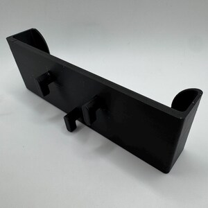 Pu&ograve; includere: Un oggetto rettangolare nero con due ganci sporgenti e bordi curvi. L'articolo sembra essere fatto di un materiale solido ed &egrave; progettato per contenere o supportare altri oggetti. Il design &egrave; semplice e funzionale.