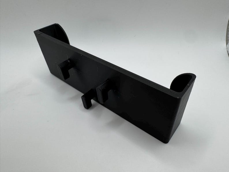 Pu&ograve; includere: Un oggetto rettangolare nero con due ganci sporgenti e bordi curvi. L'articolo sembra essere fatto di un materiale solido ed &egrave; progettato per contenere o supportare altri oggetti. Il design &egrave; semplice e funzionale.