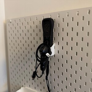 May include: A black Wii remote and nunchuck controller are mounted on a white pegboard. The controllers are connected by a black cable. The pegboard has a grid of evenly spaced holes.