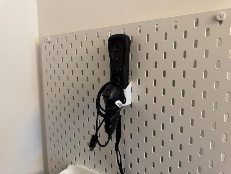 May include: A black Wii remote and nunchuck controller are mounted on a white pegboard. The controllers are connected by a black cable. The pegboard has a grid of evenly spaced holes.