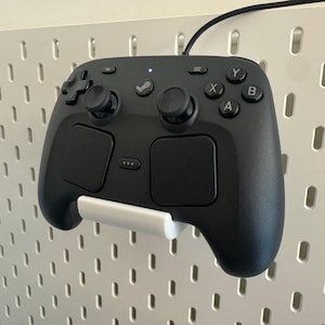 Peut inclure: Une manette Steam noire est fixée sur un panneau perforé blanc. La manette possède deux pavés tactiles, une croix directionnelle et des boutons étiquetés A, B, X et Y. Un support blanc maintient la manette. Un câble noir part du haut.