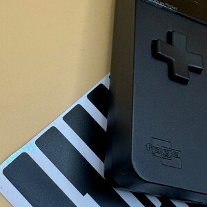May include: A black handheld gaming device with a green button and a black and white striped background. The device has a screen that displays the text "Analogue Pocket" and "FPGA".