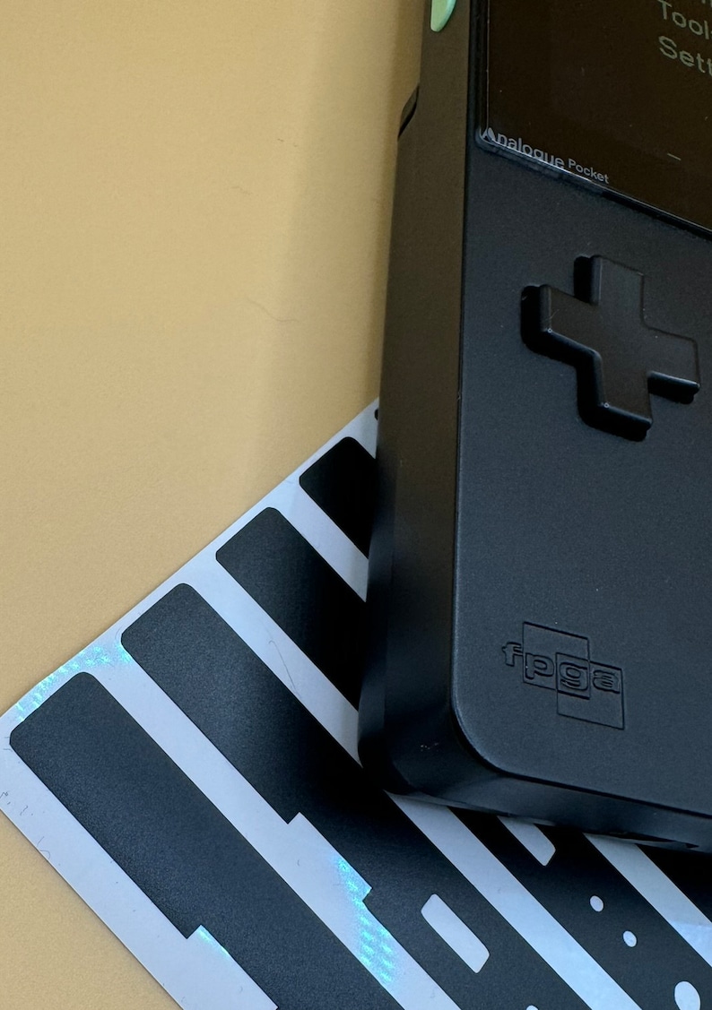 May include: A black handheld gaming device with a green button and a black and white striped background. The device has a screen that displays the text "Analogue Pocket" and "FPGA".