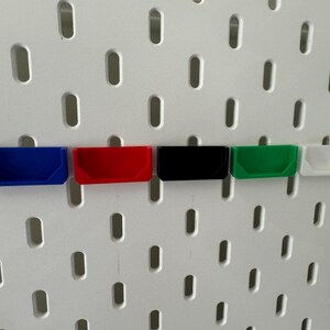 May include: Five small plastic storage bins in different colours, mounted on a white pegboard wall. The bins are blue, red, black, green, and white.