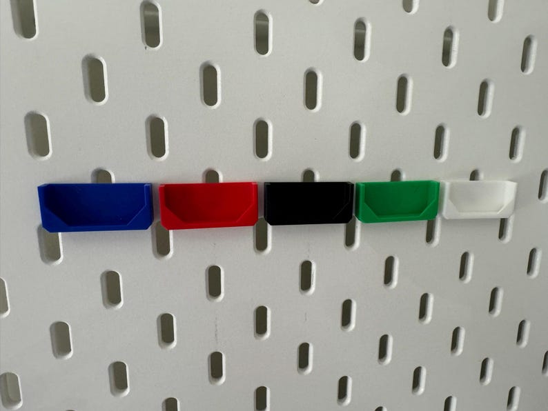 May include: Five small plastic storage bins in different colours, mounted on a white pegboard wall. The bins are blue, red, black, green, and white.