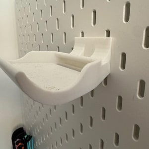 May include: A white, 3D-printed pegboard shelf with a curved design. The shelf is mounted on a white pegboard wall, showcasing its storage capabilities. The shelf is designed to hold small items.