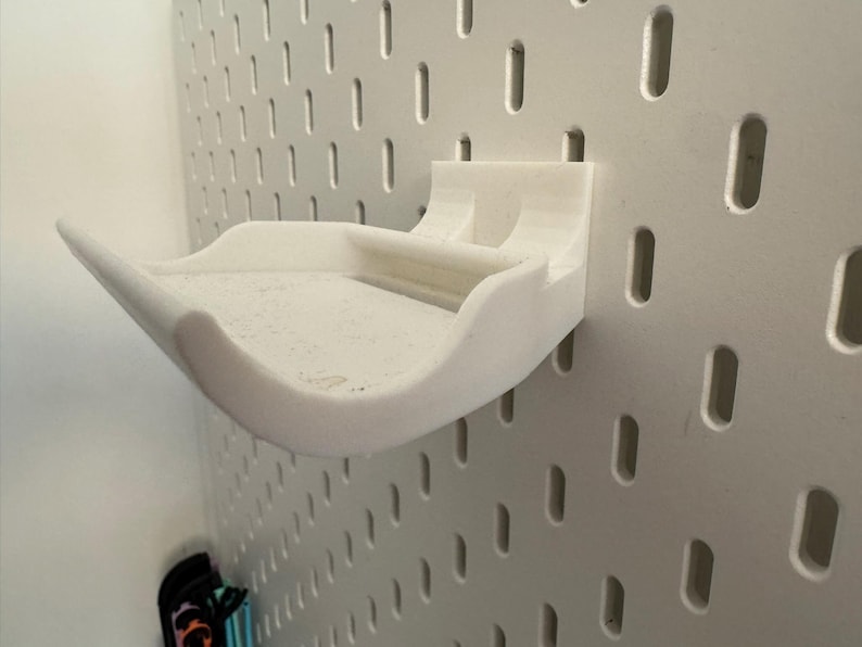 May include: A white, 3D-printed pegboard shelf with a curved design. The shelf is mounted on a white pegboard wall, showcasing its storage capabilities. The shelf is designed to hold small items.