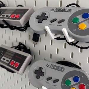 Puede incluir: Tres mandos de videojuegos montados en una pared de tablero de clavijas blanco. El mando superior es un mando Super Nintendo gris con el texto "SUPER NINTENDO" impreso en &eacute;l. El mando del medio es un mando gris de Nintendo Entertainment System. El mando inferior es otro mando Super Nintendo gris con el texto "SUPER NINTENDO" impreso en &eacute;l.