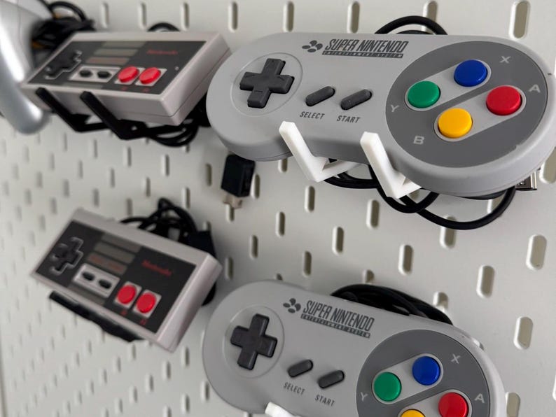 Puede incluir: Tres mandos de videojuegos montados en una pared de tablero de clavijas blanco. El mando superior es un mando Super Nintendo gris con el texto "SUPER NINTENDO" impreso en &eacute;l. El mando del medio es un mando gris de Nintendo Entertainment System. El mando inferior es otro mando Super Nintendo gris con el texto "SUPER NINTENDO" impreso en &eacute;l.