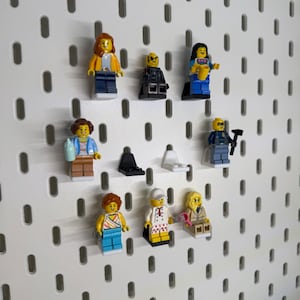 K&ouml;nnte beinhalten: Eine Sammlung von Lego-Minifiguren, jede mit einzigartigen Outfits und Accessoires, auf einer wei&szlig;en Lochwand ausgestellt. Die Minifiguren zeigen Charaktere in verschiedenen Outfits, wie einer gelben Jacke, einem schwarzen Anzug und einem blauen Kleid, die eine Vielzahl von Stilen zeigen.