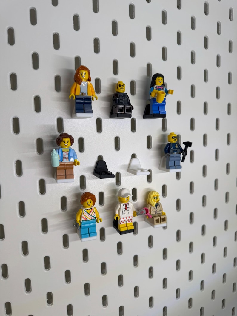 K&ouml;nnte beinhalten: Eine Sammlung von Lego-Minifiguren, jede mit einzigartigen Outfits und Accessoires, auf einer wei&szlig;en Lochwand ausgestellt. Die Minifiguren zeigen Charaktere in verschiedenen Outfits, wie einer gelben Jacke, einem schwarzen Anzug und einem blauen Kleid, die eine Vielzahl von Stilen zeigen.