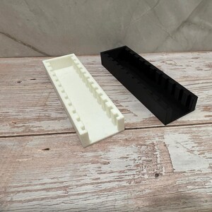 Puede incluir: Dos contenedores de almacenamiento rectangulares, uno blanco y otro negro. Ambos tienen la parte superior abierta y divisores internos. Est&aacute;n colocados sobre una superficie de madera con un patr&oacute;n marr&oacute;n claro y blanco.