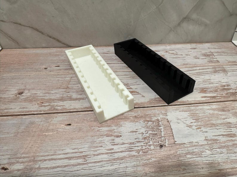 Puede incluir: Dos contenedores de almacenamiento rectangulares, uno blanco y otro negro. Ambos tienen la parte superior abierta y divisores internos. Est&aacute;n colocados sobre una superficie de madera con un patr&oacute;n marr&oacute;n claro y blanco.