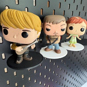Op de afbeelding: Drie popcultuurfiguurtjes die op zwarte en witte planken worden getoond. De figuurtjes zijn van een blonde man met een luit, een man met bruin haar met een kruisboog en een roodharige vrouw met een jurk.