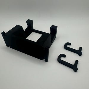 K&ouml;nnte beinhalten: Eine schwarze Wandhalterung aus Kunststoff mit zwei Haken. Die Halterung hat eine rechteckige Form mit einer quadratischen &Ouml;ffnung in der Mitte. Die Haken sind J-f&ouml;rmig.