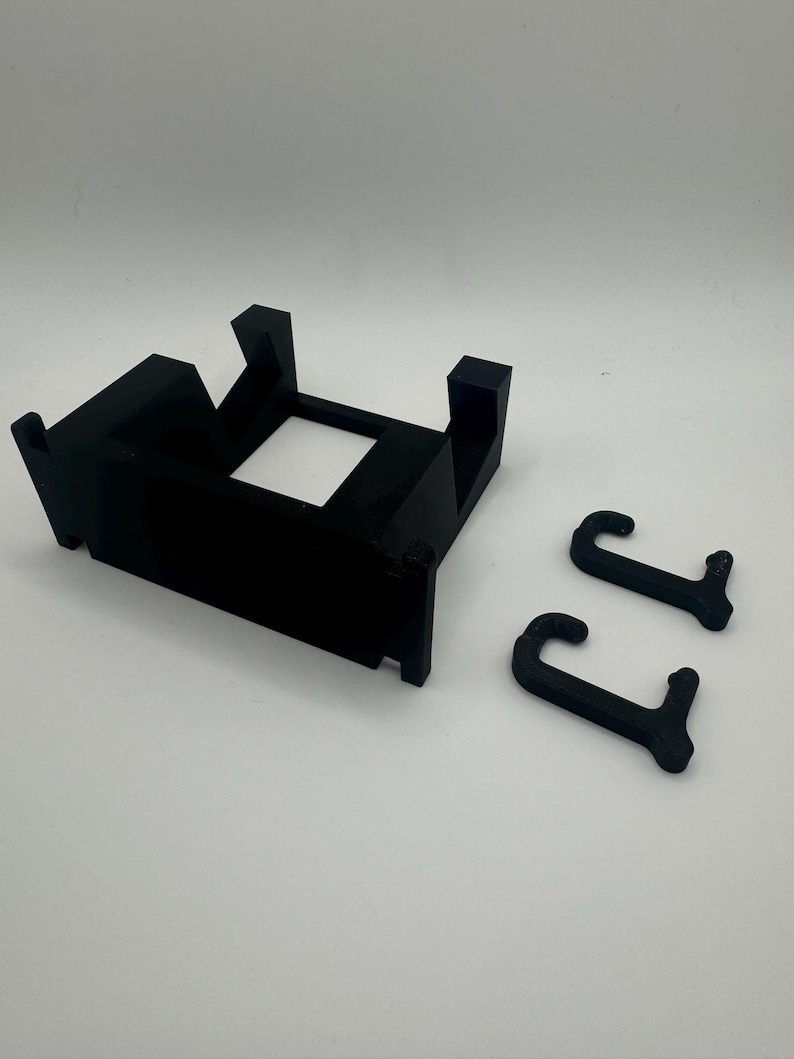 K&ouml;nnte beinhalten: Eine schwarze Wandhalterung aus Kunststoff mit zwei Haken. Die Halterung hat eine rechteckige Form mit einer quadratischen &Ouml;ffnung in der Mitte. Die Haken sind J-f&ouml;rmig.