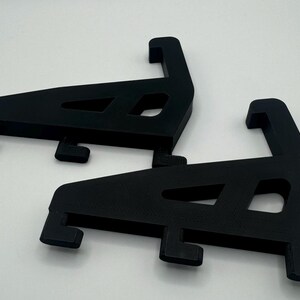 May include: Two black plastic shelf brackets, each with a triangular shape and a curved lip on the top. The brackets are designed to support shelves.