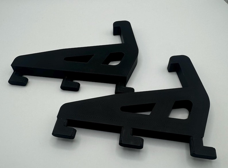 May include: Two black plastic shelf brackets, each with a triangular shape and a curved lip on the top. The brackets are designed to support shelves.