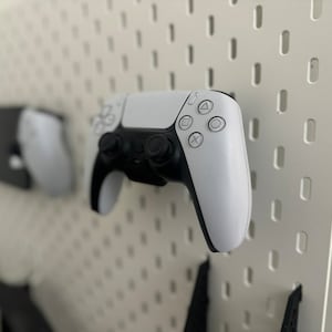 May include: Two white wireless game controllers are mounted on a white pegboard wall. The controllers have black buttons and a black PlayStation logo.