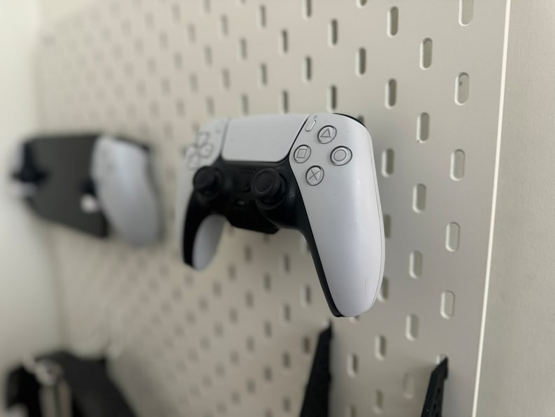 May include: Two white wireless game controllers are mounted on a white pegboard wall. The controllers have black buttons and a black PlayStation logo.