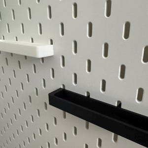 May include: A white pegboard with a series of evenly spaced, oblong holes. Two rectangular shelves, one white and one black, are inserted into the board. A green object is visible in the upper left corner.