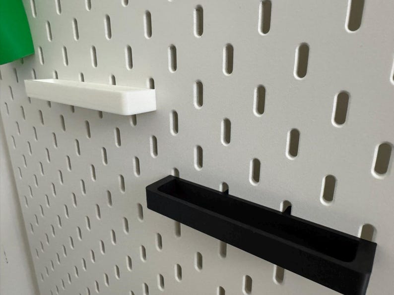 May include: A white pegboard with a series of evenly spaced, oblong holes. Two rectangular shelves, one white and one black, are inserted into the board. A green object is visible in the upper left corner.