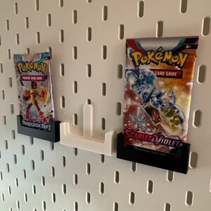 Op de afbeelding: Twee Pok&eacute;mon trading card game booster packs weergegeven op een wit pegboard. De packs worden vastgehouden in zwarte en witte 3D-geprinte houders. E&eacute;n pack is gelabeld "Paradox Rift", de andere "Scarlet & Violet".