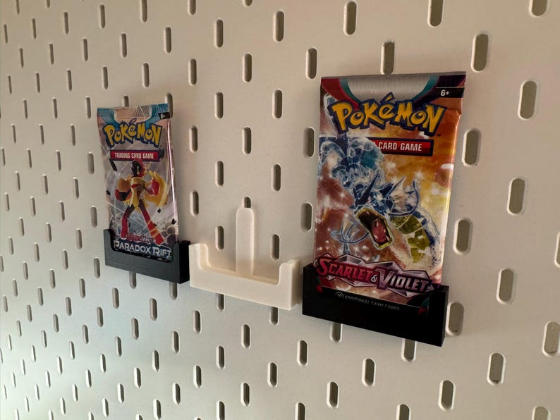 Op de afbeelding: Twee Pok&eacute;mon trading card game booster packs weergegeven op een wit pegboard. De packs worden vastgehouden in zwarte en witte 3D-geprinte houders. E&eacute;n pack is gelabeld "Paradox Rift", de andere "Scarlet & Violet".