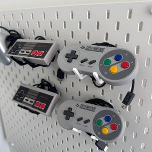 Puede incluir: Tres mandos de videojuegos colgados en una pared de tablero de clavijas. Dos mandos grises de Super Nintendo con el texto "SUPER NINTENDO" y un mando gris de Nintendo Entertainment System.
