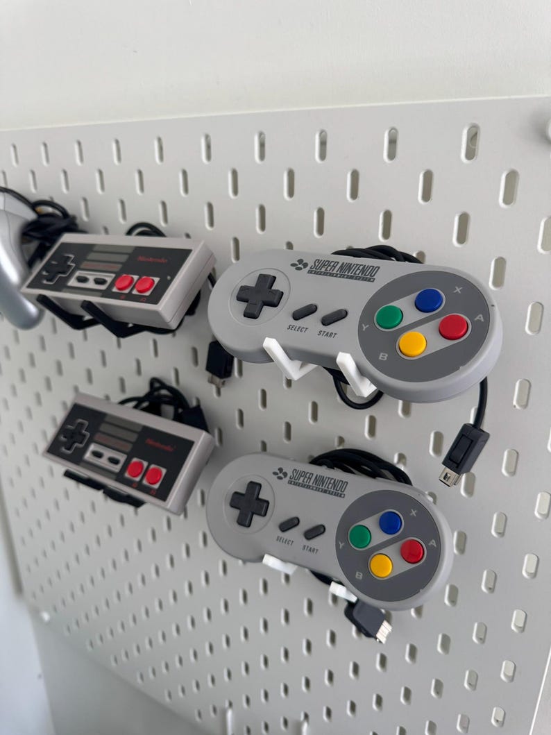 Puede incluir: Tres mandos de videojuegos colgados en una pared de tablero de clavijas. Dos mandos grises de Super Nintendo con el texto "SUPER NINTENDO" y un mando gris de Nintendo Entertainment System.
