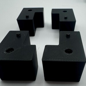 May include: Four black plastic corner brackets with a square shape and a small hole in the centre of each side.