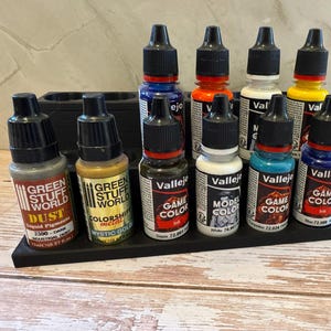 Puede incluir: Una colecci&oacute;n de peque&ntilde;os botes de pintura de varios colores, incluyendo azul, naranja, blanco y amarillo, est&aacute;n dispuestos en un organizador negro. Los botes tienen tapas negras y etiquetas con texto, incluyendo "Vallejo" y "Game Color".