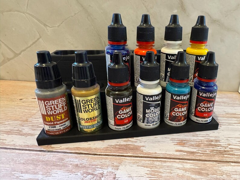 Puede incluir: Una colecci&oacute;n de peque&ntilde;os botes de pintura de varios colores, incluyendo azul, naranja, blanco y amarillo, est&aacute;n dispuestos en un organizador negro. Los botes tienen tapas negras y etiquetas con texto, incluyendo "Vallejo" y "Game Color".