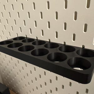 May include: A black, rectangular pegboard organizer with 12 circular holes. The organizer is designed to hold small items and is mounted on a white pegboard wall. The product is made of a solid, matte material.