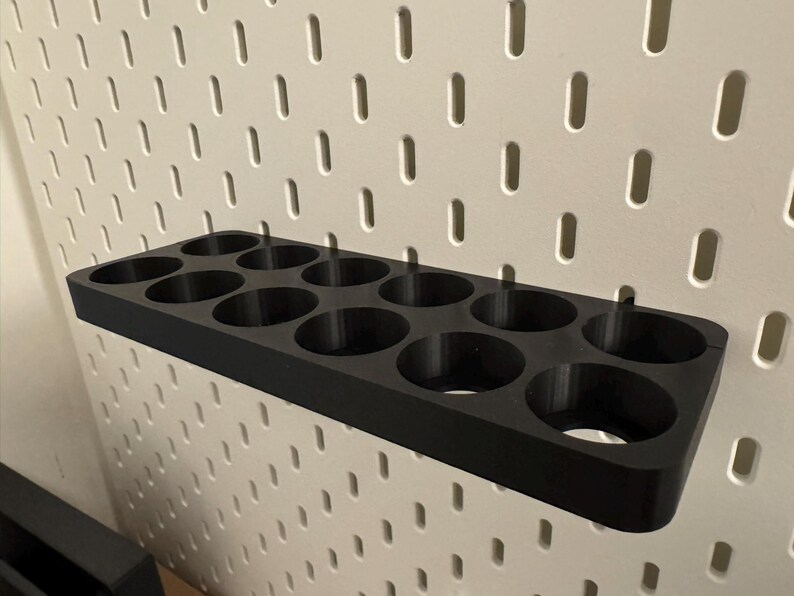 May include: A black, rectangular pegboard organizer with 12 circular holes. The organizer is designed to hold small items and is mounted on a white pegboard wall. The product is made of a solid, matte material.