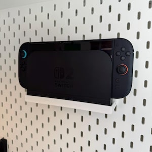 Puede incluir: Una consola Nintendo Switch negra acoplada en un estante blanco, montada en una pared perforada blanca. La consola tiene una pantalla y mandos adjuntos con detalles en azul y rojo. El logotipo de Nintendo Switch es visible.