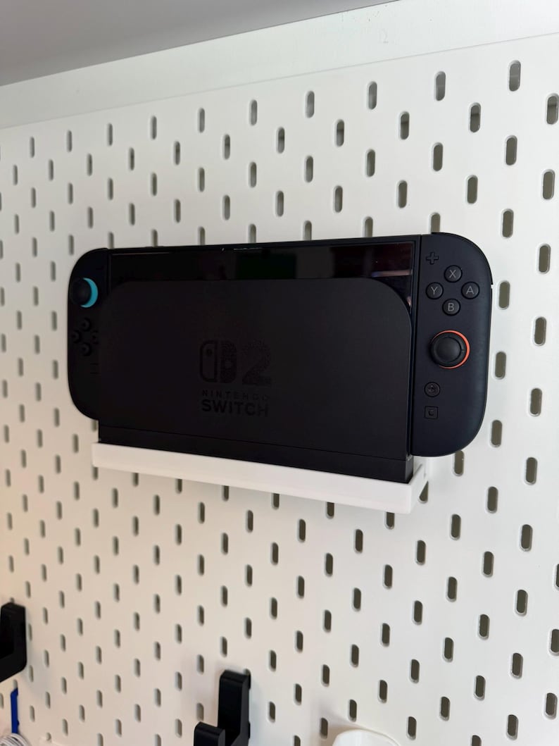 Puede incluir: Una consola Nintendo Switch negra acoplada en un estante blanco, montada en una pared perforada blanca. La consola tiene una pantalla y mandos adjuntos con detalles en azul y rojo. El logotipo de Nintendo Switch es visible.