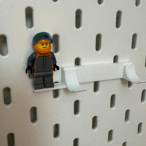 May include: A white plastic pegboard shelf with a small, grey and orange Lego minifigure standing on it.
