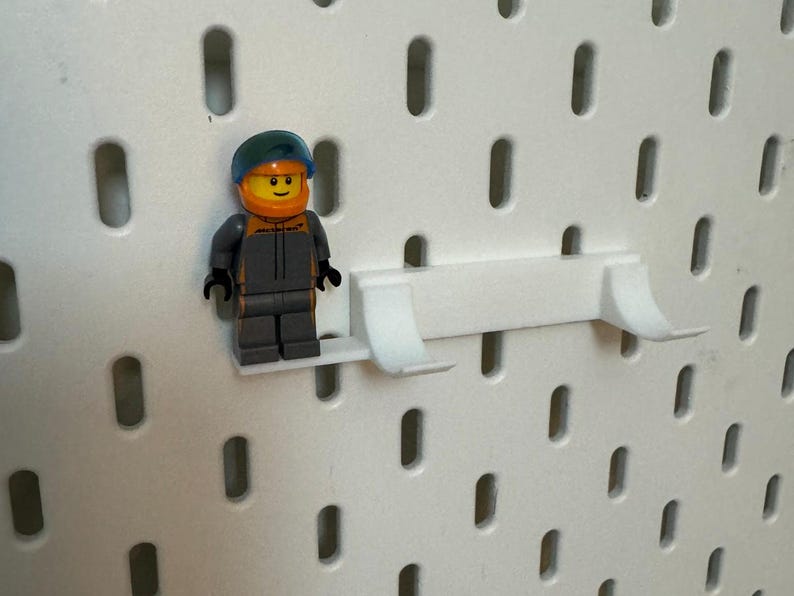 May include: A white plastic pegboard shelf with a small, grey and orange Lego minifigure standing on it.