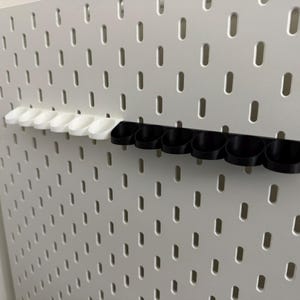 以下が含まれることがあります： 白のペグボードに、2列の収納カップが取り付けられています。1列は白いカップ、もう1列は黒いカップです。ペグボードには、等間隔に配置された楕円形の穴があります。