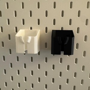 May include: Two 3D-printed storage containers. One is white, the other is black. Both have a rectangular shape with a slot in the front. They are designed to attach to a pegboard. The pegboard has a white surface with evenly spaced holes.