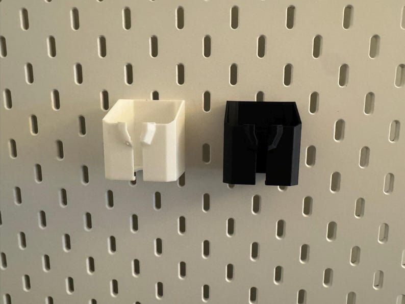 May include: Two 3D-printed storage containers. One is white, the other is black. Both have a rectangular shape with a slot in the front. They are designed to attach to a pegboard. The pegboard has a white surface with evenly spaced holes.