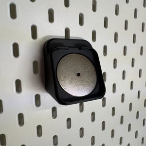 May include: A black, square pegboard accessory with a circular, silver magnet. It's mounted on a white pegboard with evenly spaced oval holes. The magnet is centrally positioned within the accessory.