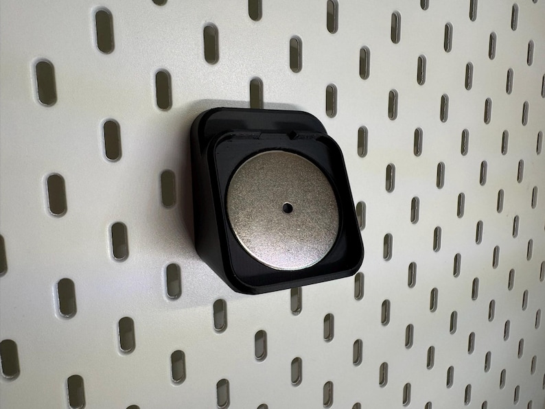 May include: A black, square pegboard accessory with a circular, silver magnet. It's mounted on a white pegboard with evenly spaced oval holes. The magnet is centrally positioned within the accessory.