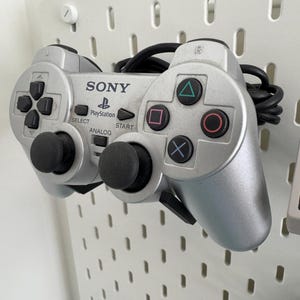 Puede incluir: Un mando de PlayStation plateado con botones negros y palancas anal&oacute;gicas. El mando tiene el logotipo de PlayStation y la palabra "SONY" impresa en &eacute;l.