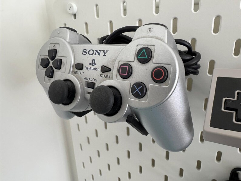 Puede incluir: Un mando de PlayStation plateado con botones negros y palancas anal&oacute;gicas. El mando tiene el logotipo de PlayStation y la palabra "SONY" impresa en &eacute;l.