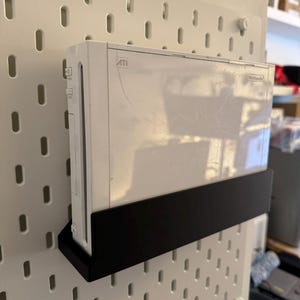 Peut inclure: Une console Nintendo Wii blanche fix&eacute;e sur une &eacute;tag&egrave;re noire, elle-m&ecirc;me fix&eacute;e &agrave; un panneau perfor&eacute; blanc. La console est rectangulaire avec le logo Nintendo. Le panneau perfor&eacute; a des trous espac&eacute;s.