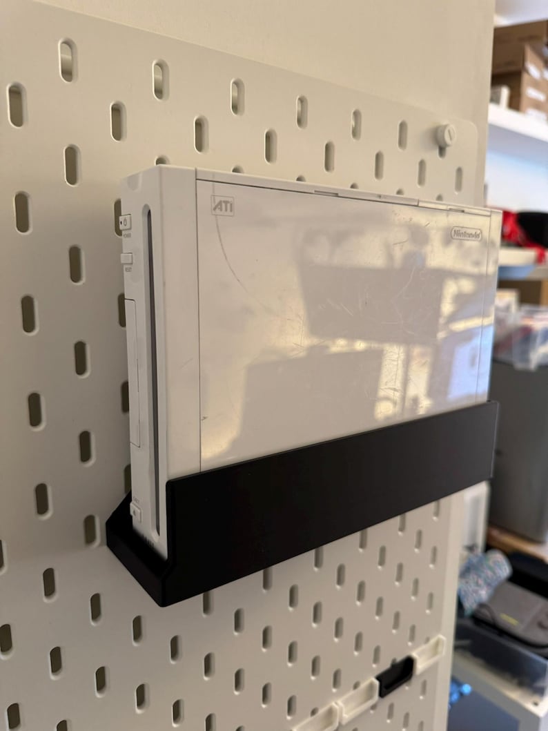 Peut inclure: Une console Nintendo Wii blanche fix&eacute;e sur une &eacute;tag&egrave;re noire, elle-m&ecirc;me fix&eacute;e &agrave; un panneau perfor&eacute; blanc. La console est rectangulaire avec le logo Nintendo. Le panneau perfor&eacute; a des trous espac&eacute;s.